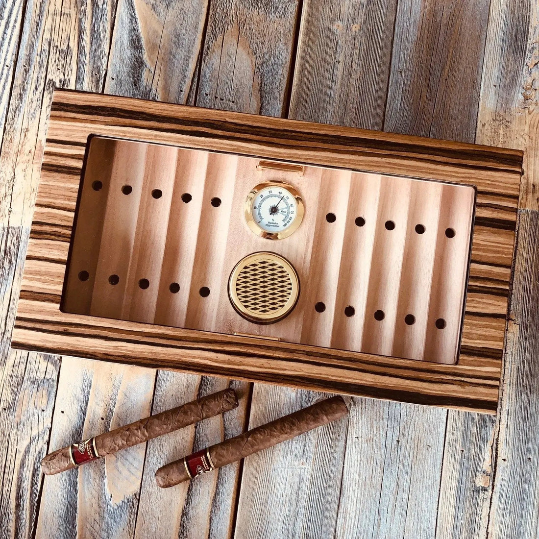 Glass top desktop cigar humidor holding a row of cigars in various vitola sizes including robustos, toros, and a full-length churchill