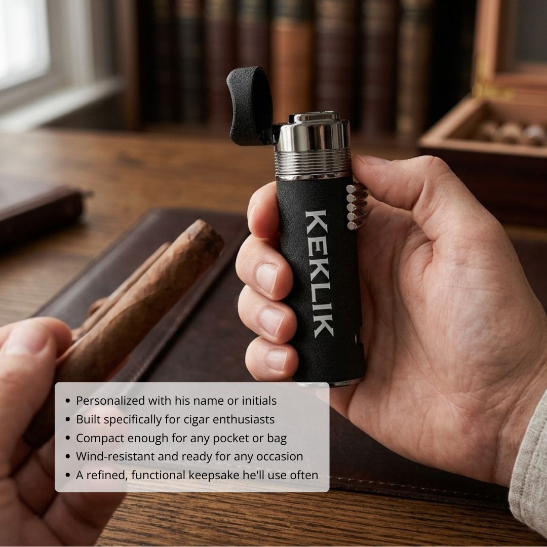 Black textured cigar lighter with sample monogram "KEKLIK" engraved on side, held in hand above wooden cigar tray, showing metal band and flip-top ignition mechanism