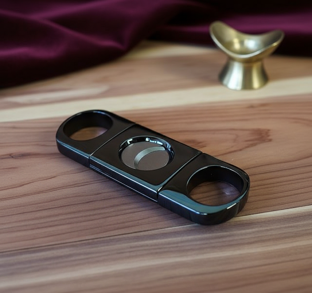 Gun metal double-blade guillotine cigar cutter with dual oval openings, displayed on wooden surface with brass rest in background