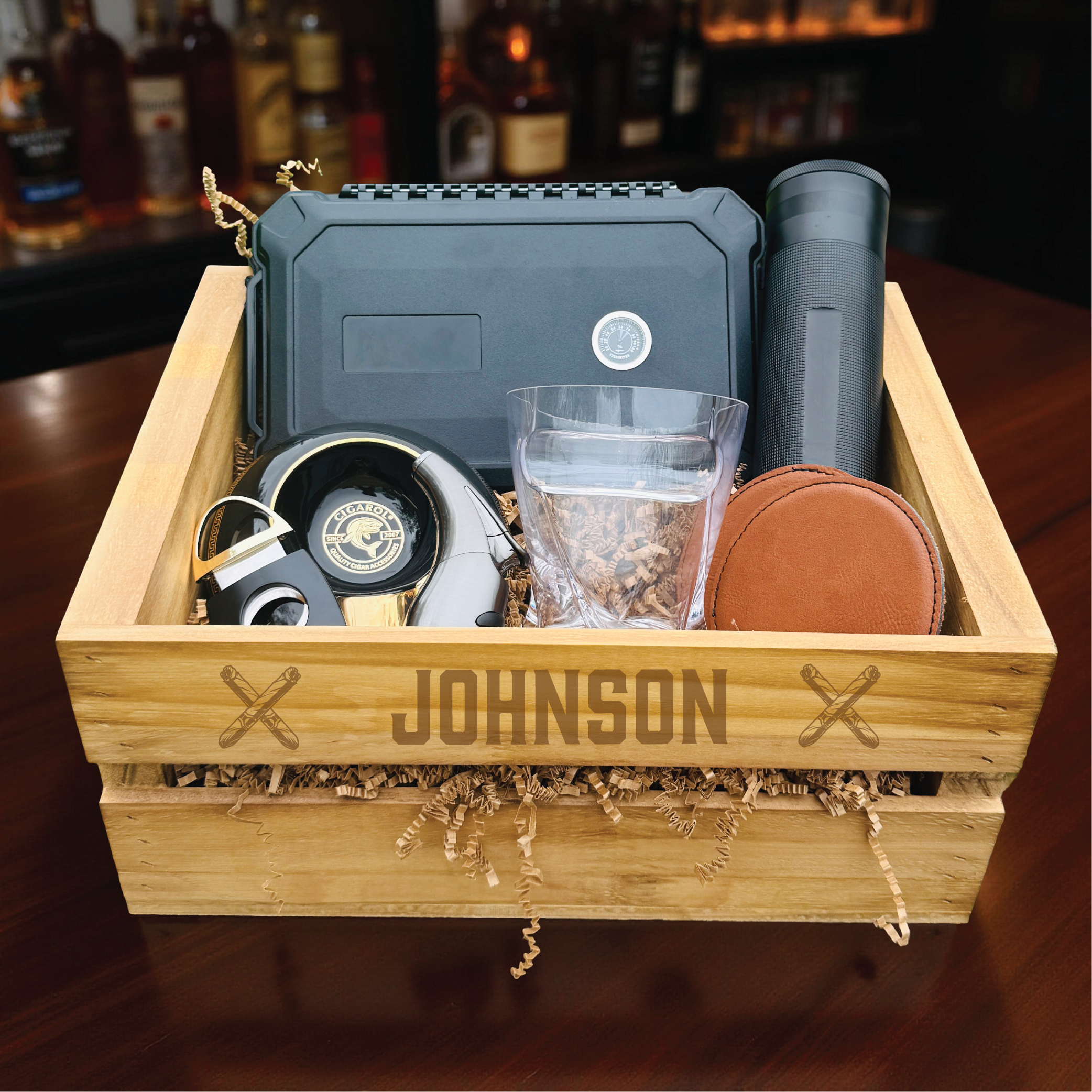 Wooden crate engraved with sample name "JOHNSON" containing cigar humidor, cigar cutter, whiskey glass, lighter, leather cigar case, and travel tube displayed on wood surface with bar backdrop.