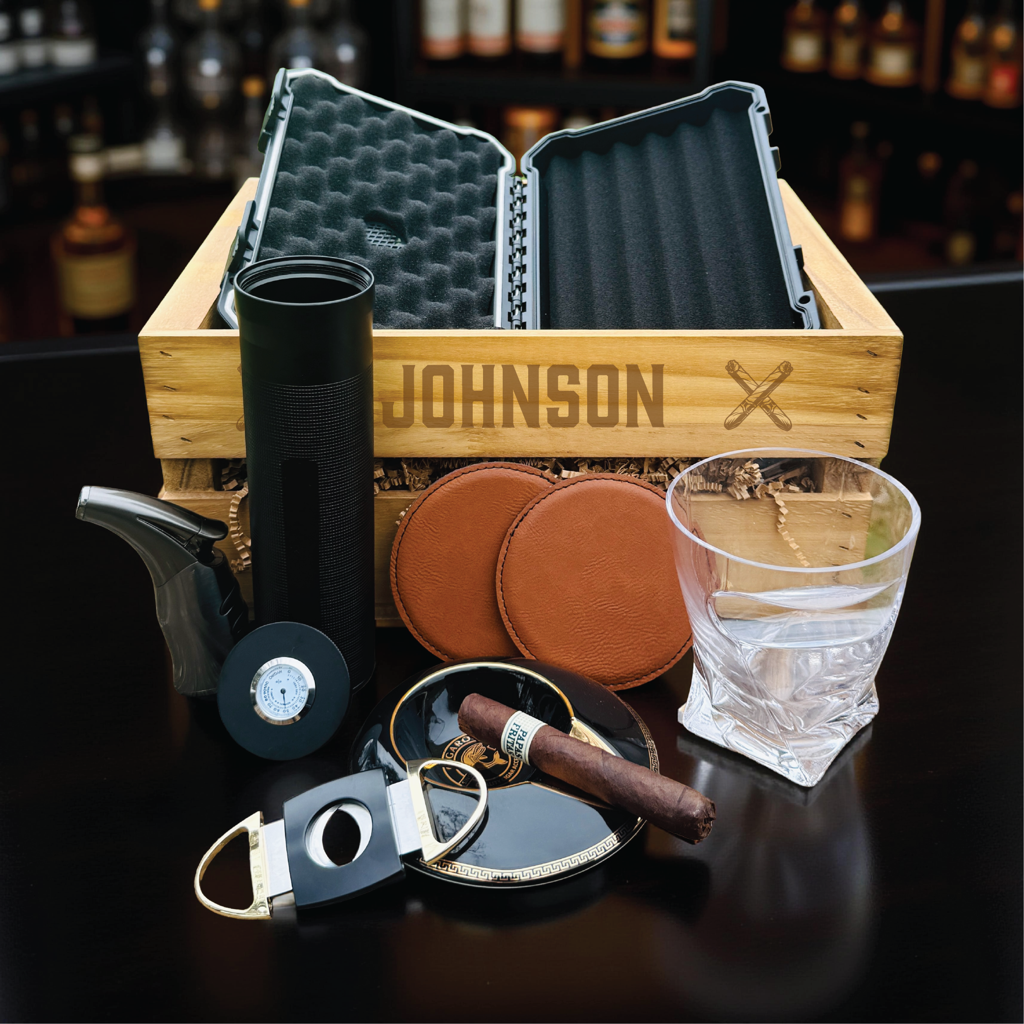 Personalized wooden crate engraved with sample name "JOHNSON" displaying open cigar travel case, black lighter, tan leather coasters, cigar cutter, ashtray, glass, and humidor accessories inside
