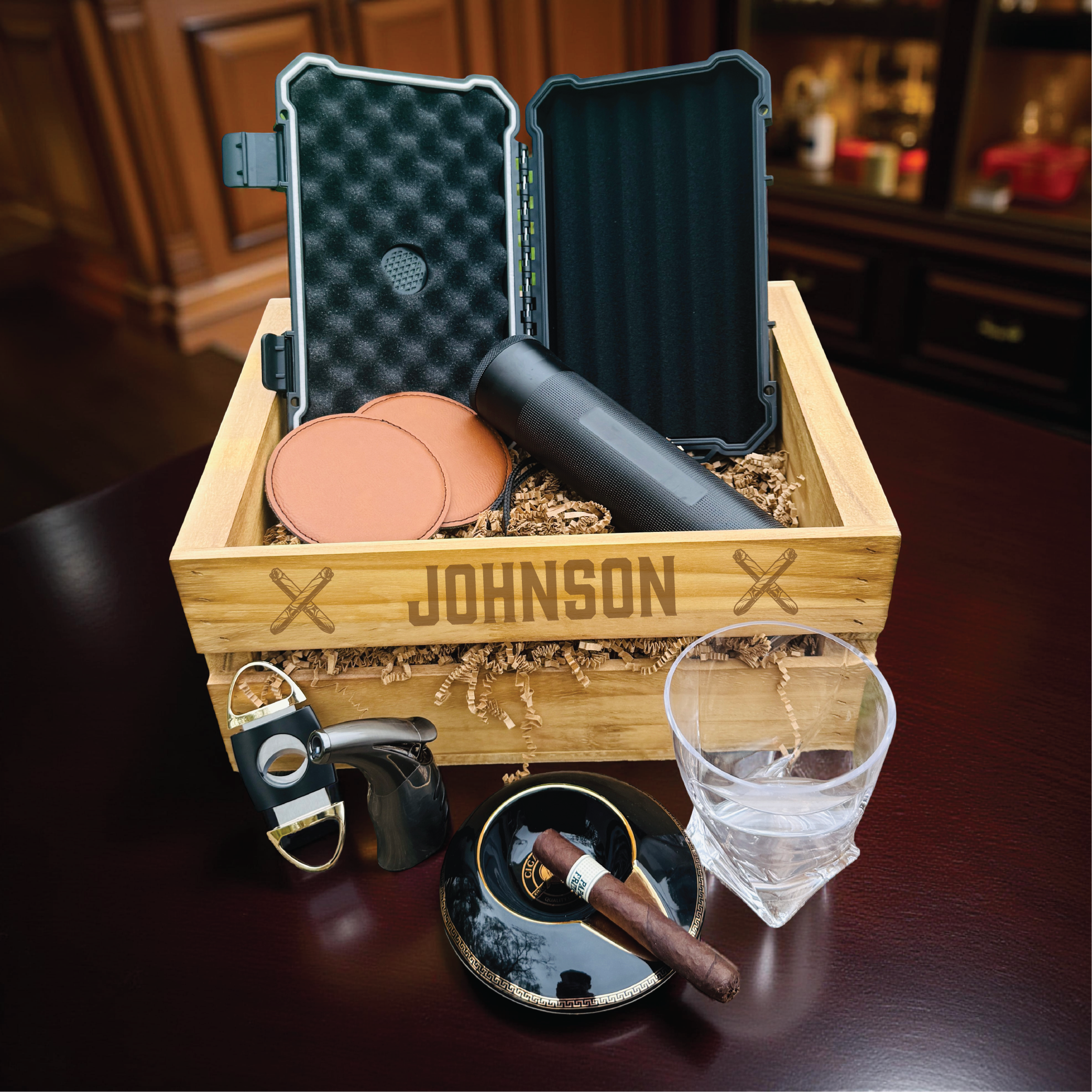 Personalized wooden crate engraved with sample name "JOHNSON" containing cigar travel case, leather cigar coasters, cutter, lighter, whiskey glass, and ashtray displayed on dark wood surface