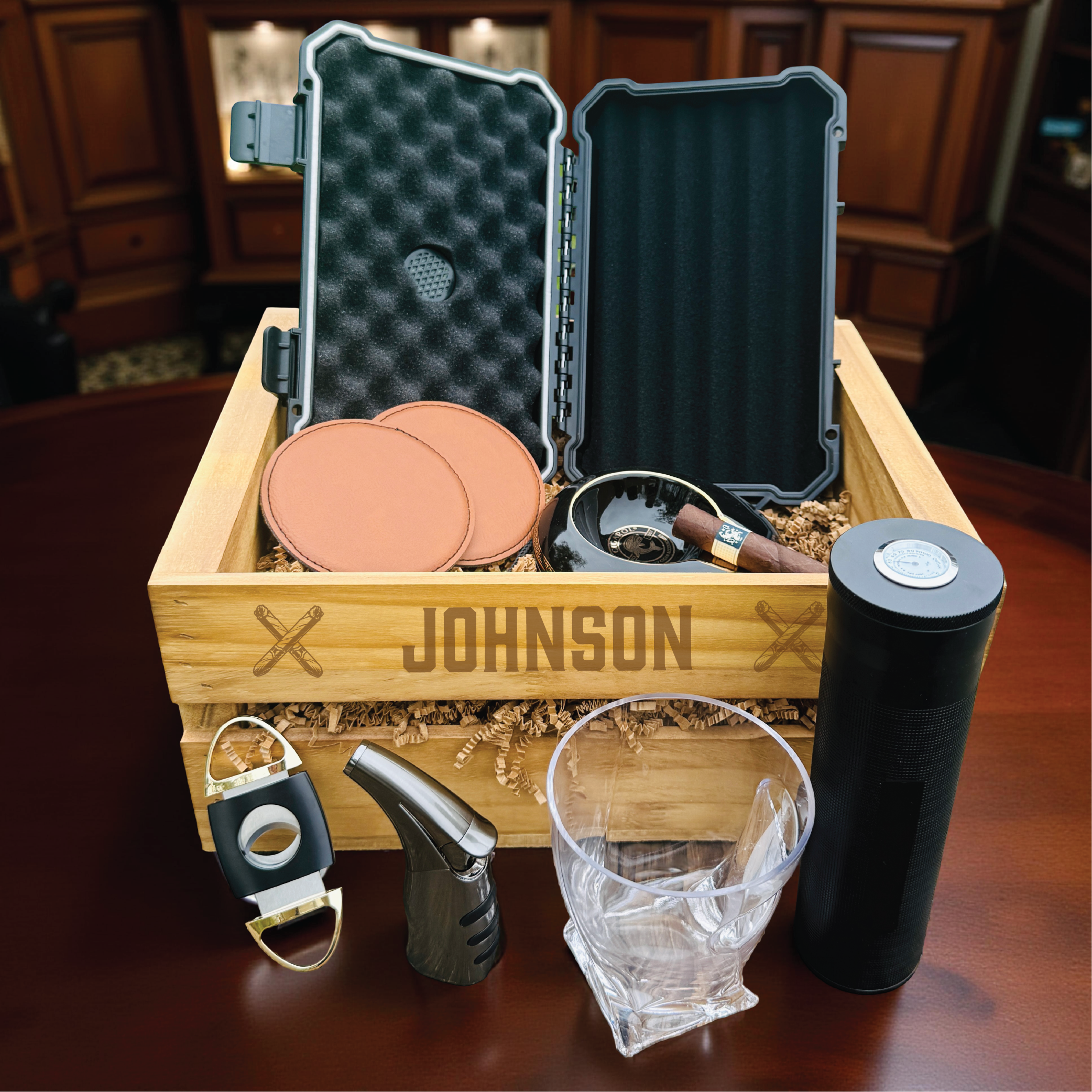 Personalized wooden crate engraved with sample name "JOHNSON" displayed open with cigar accessories including humidor, cutters, lighters, ashtrays, travel case, and whiskey glass