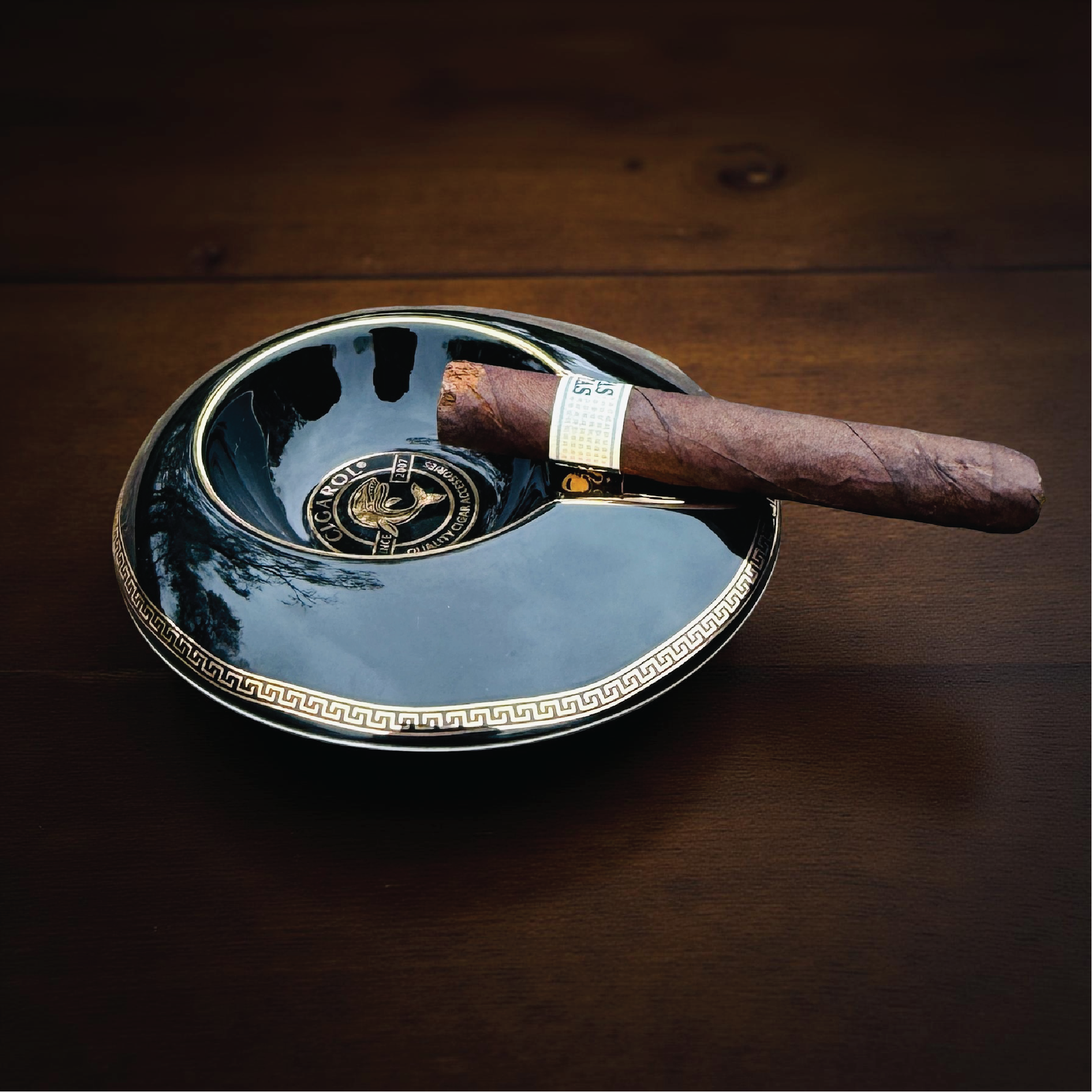 Black ceramic cigar ashtray with gold Greek key trim and built-in cigar rest, holding a lit cigar on a wooden surface