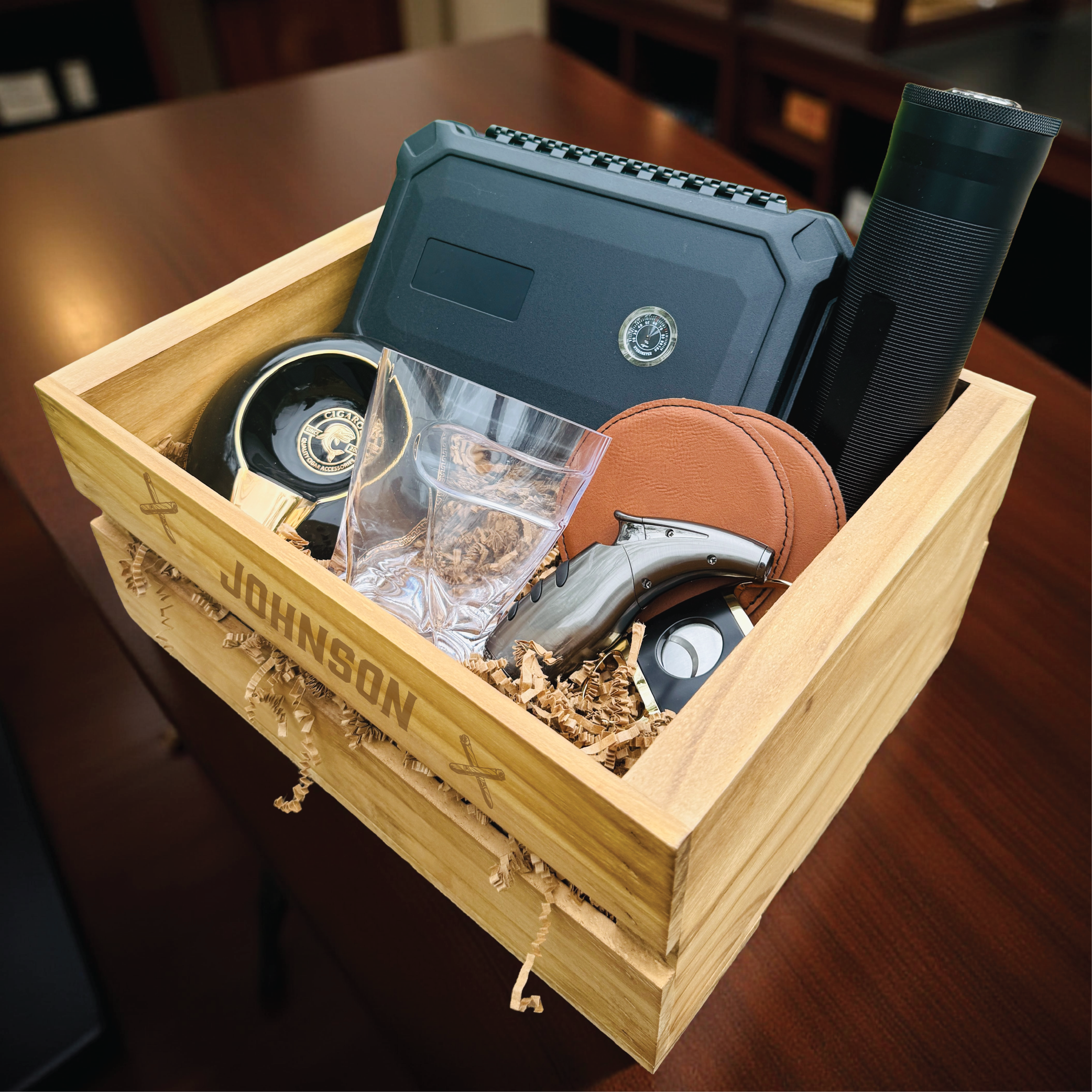 Open wooden crate engraved with sample name "JOHNSON" containing blue humidor, crystal whiskey glass, tan leather cigar case, black lighter, ashtray, and decorative filler