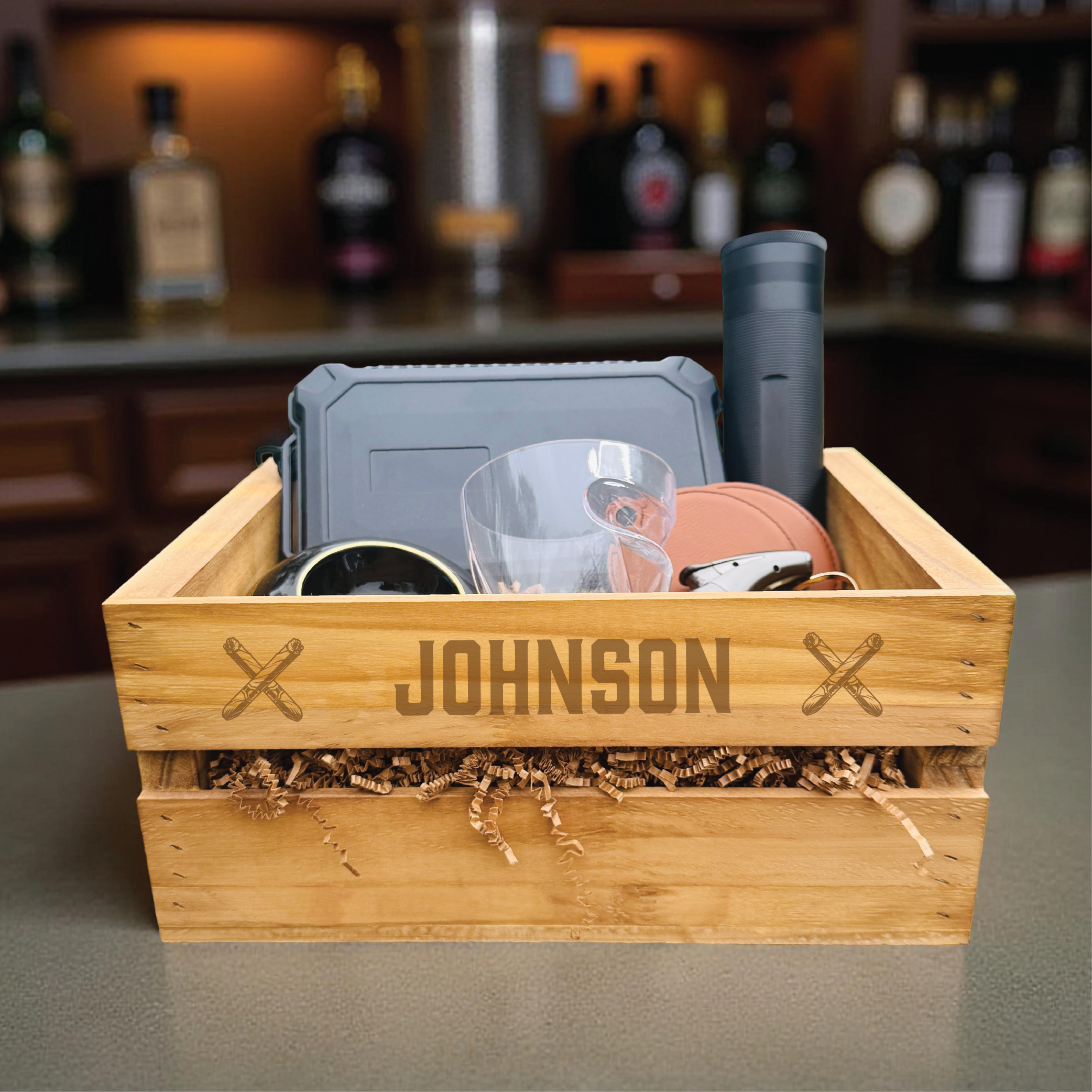 Wooden crate laser-engraved with sample name "JOHNSON" and decorative crossed cigar symbols, filled with gray humidor, whiskey glass, black lighter, coral leather cigar case, and natural wood shavings