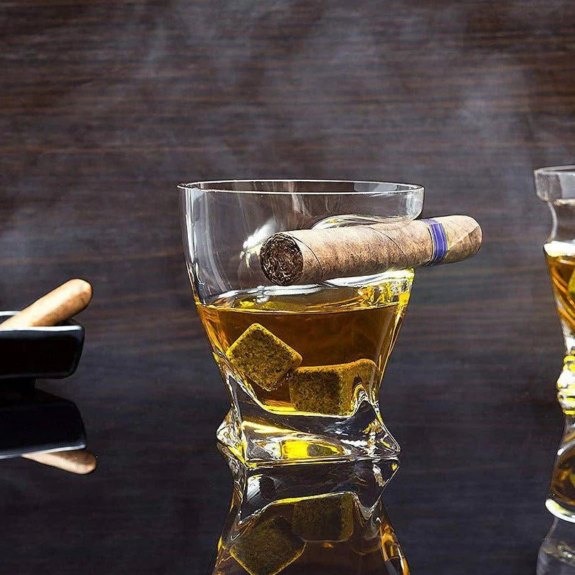 Whiskey glass with built-in cigar rest holding a lit cigar and ice cubes, displayed on dark wood surface alongside smoking accessories
